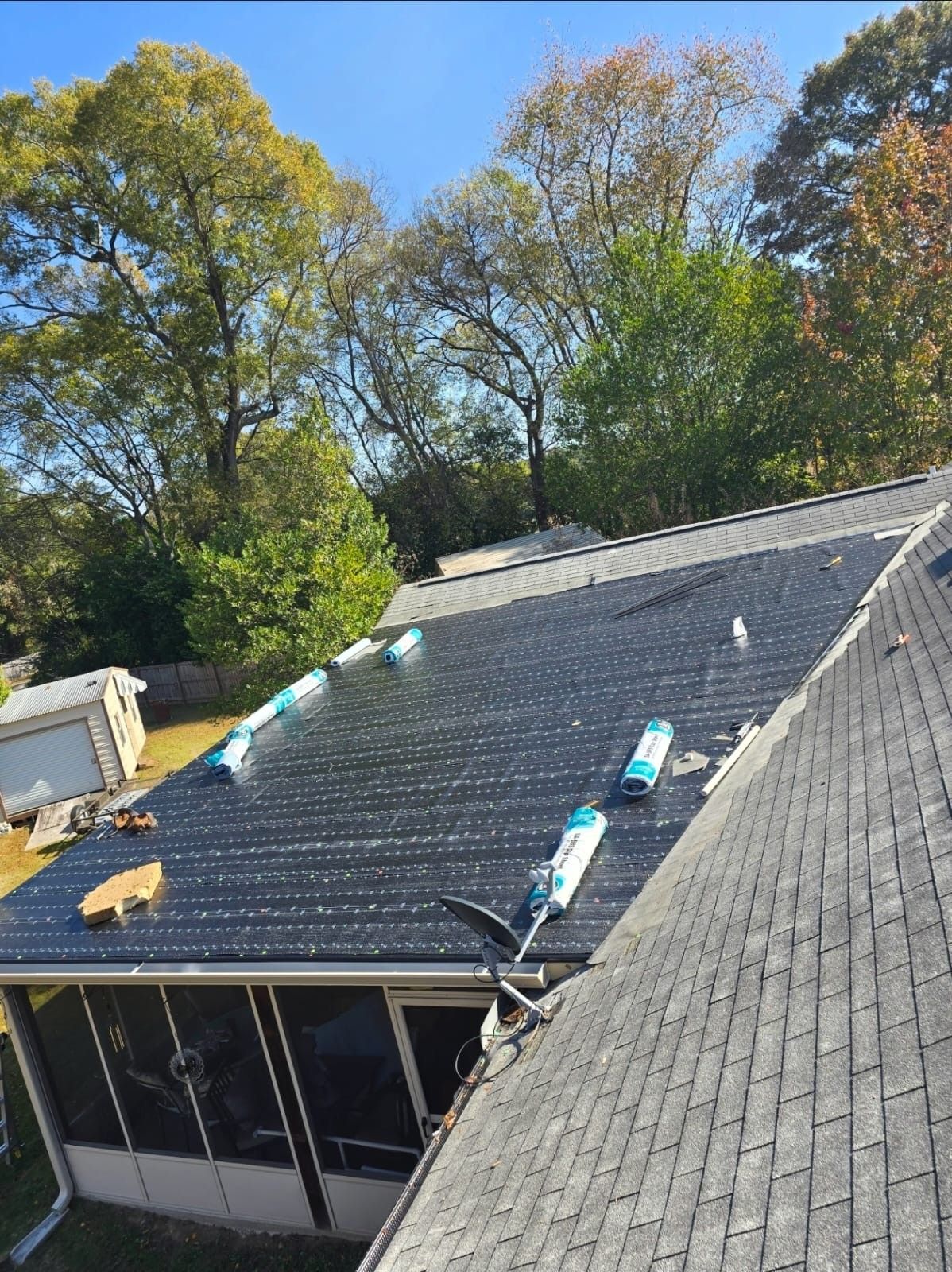Solar panels installed on a dark roof, with a screen porch below and trees in the background on a sunny day.