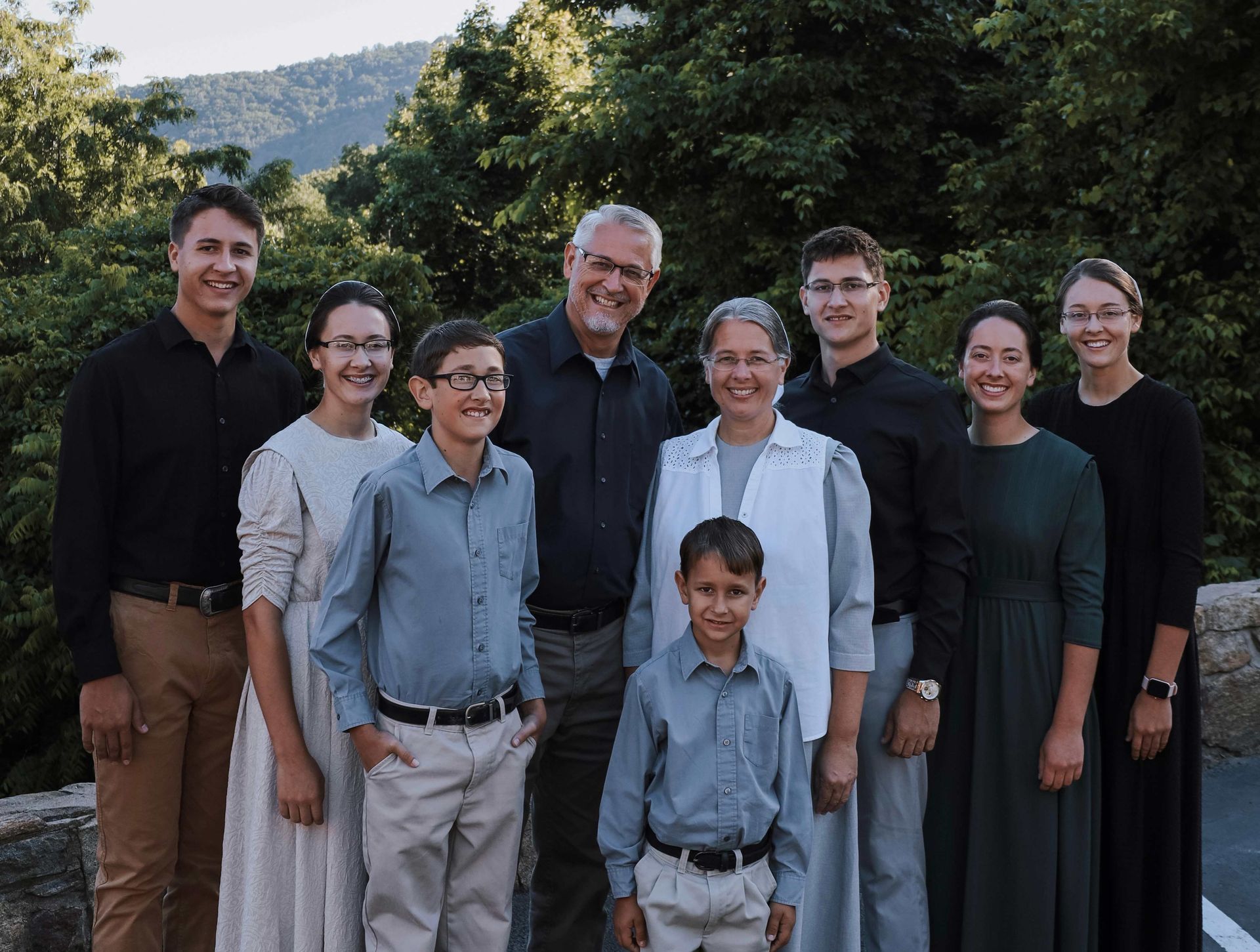 Family group posing outside with mountain backdrop. Adults and children wear conservative clothing.