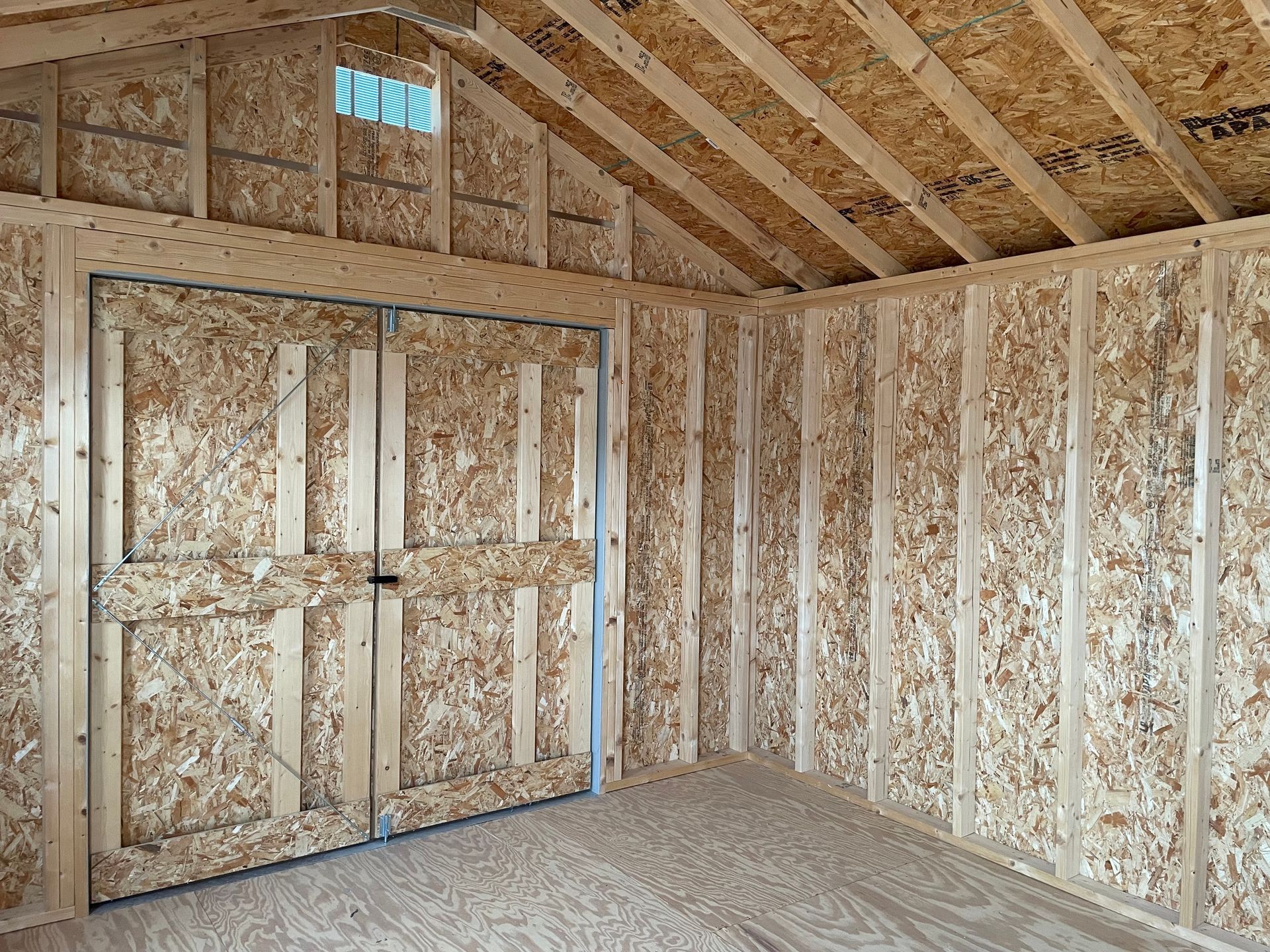 Interior of a wooden shed with double doors, oriented strand board walls and floor, and an overhead vent.
