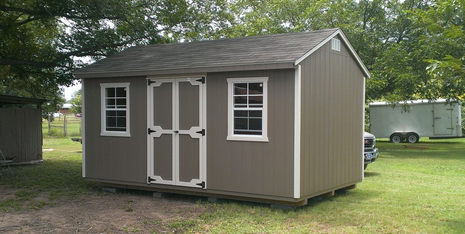 Lawn with wooden fence, shed, and a brick house. Sun shining through tree branches on a sunny day.