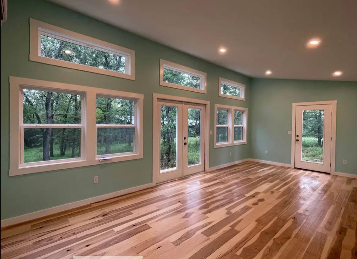 A light-filled room with wood floors, light blue walls, and large windows overlooking a wooded area.
