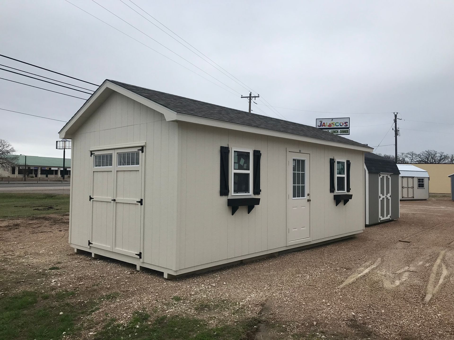 Tan shed with black shutters, white door, and windows, on gravel.