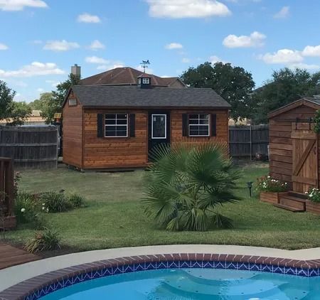 Wooden shed in a backyard with a pool, palm tree, and blue sky.