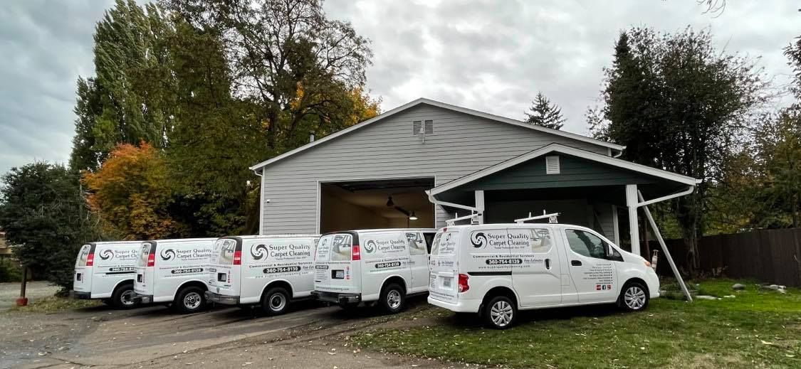 Four white vans parked in front of a garage with a green roof under an overcast sky.