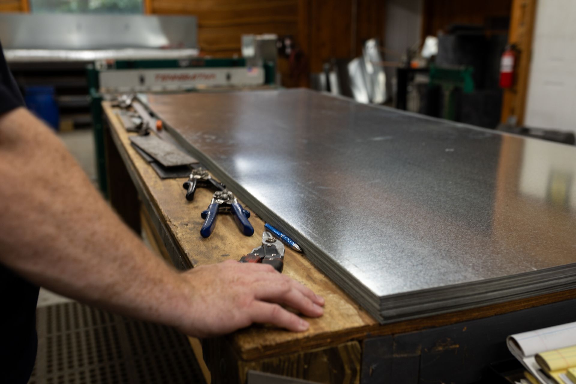 Man's hand resting on a stack of galvanized steel sheets on a workbench, with tools and machinery in the background.