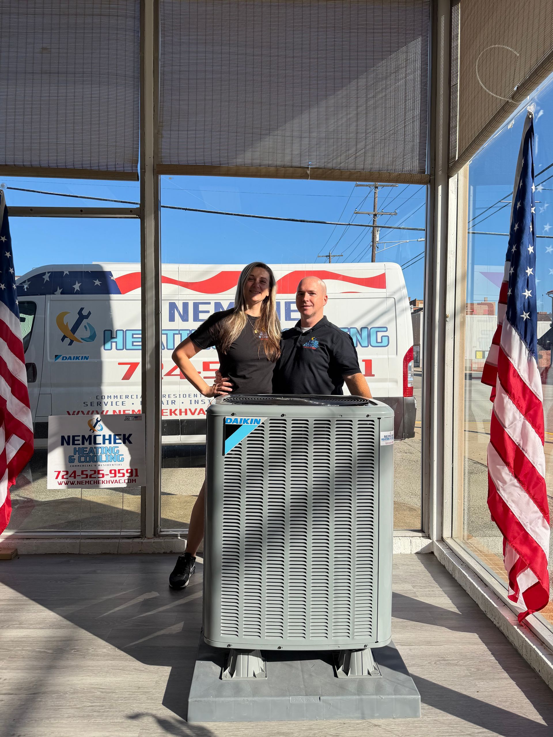 Two people stand at a podium on a sunny stage with American flags and a truck in the background.