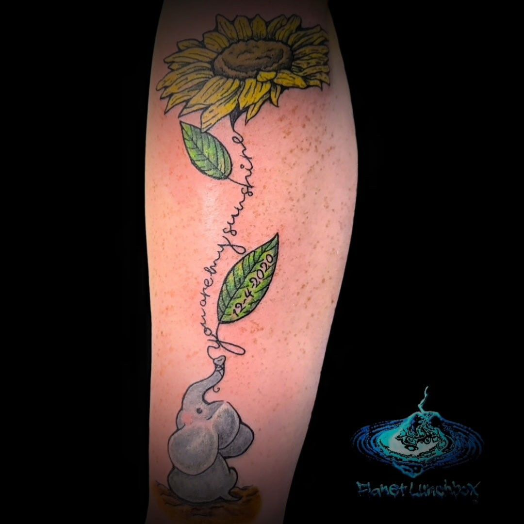 Sunflower tattoo with a vine forming an elephant shape and two green leaves on a forearm