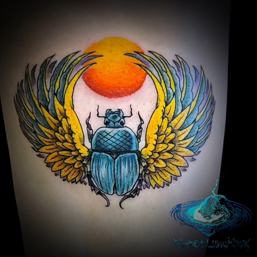 Blue scarab beetle with gold wings beneath an orange sun, tattoo-style design on skin