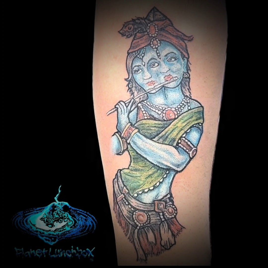 Blue-skinned tattooed figure holding a flute, with green sari and floral headpiece on a forearm