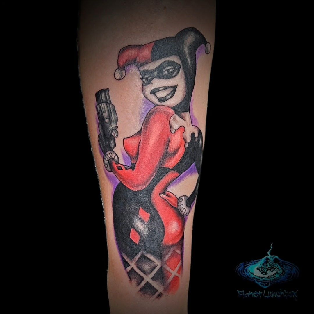 Colorful tattoo of Harley Quinn holding a gun, wearing a red-and-black jester outfit.
