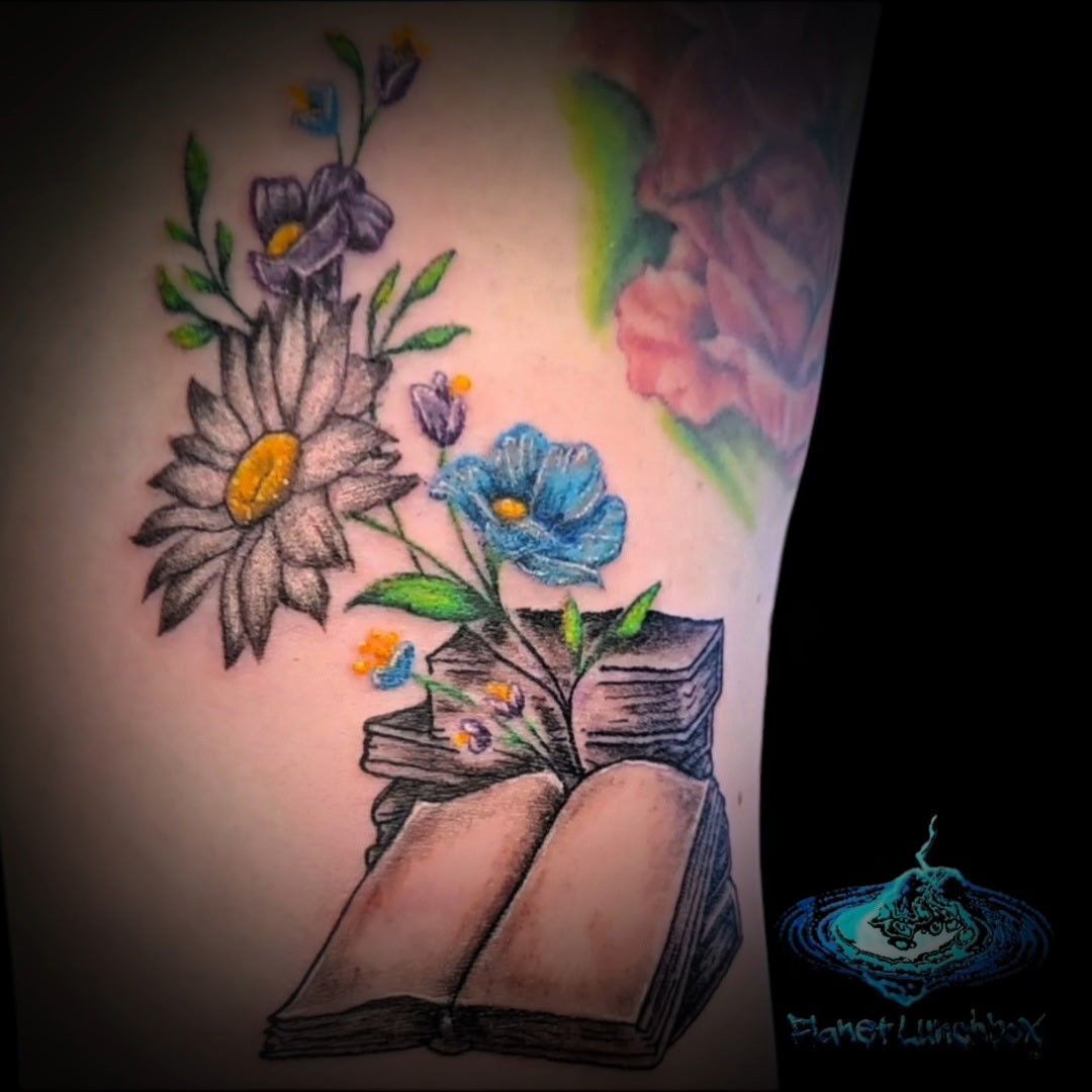 Colorful floral tattoo on skin: daisies, blue rose, open book, and pink flower with soft watercolor shading