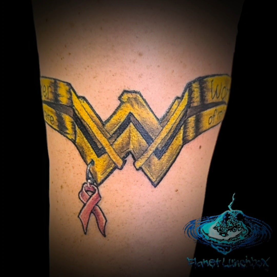 Wonder Woman logo tattoo on skin with a small pink ribbon hanging from the left side
