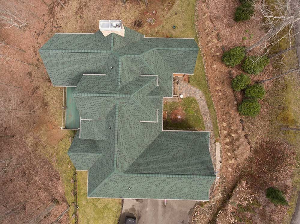 Aerial view of a house with a green shingle roof, surrounded by brown and green foliage.