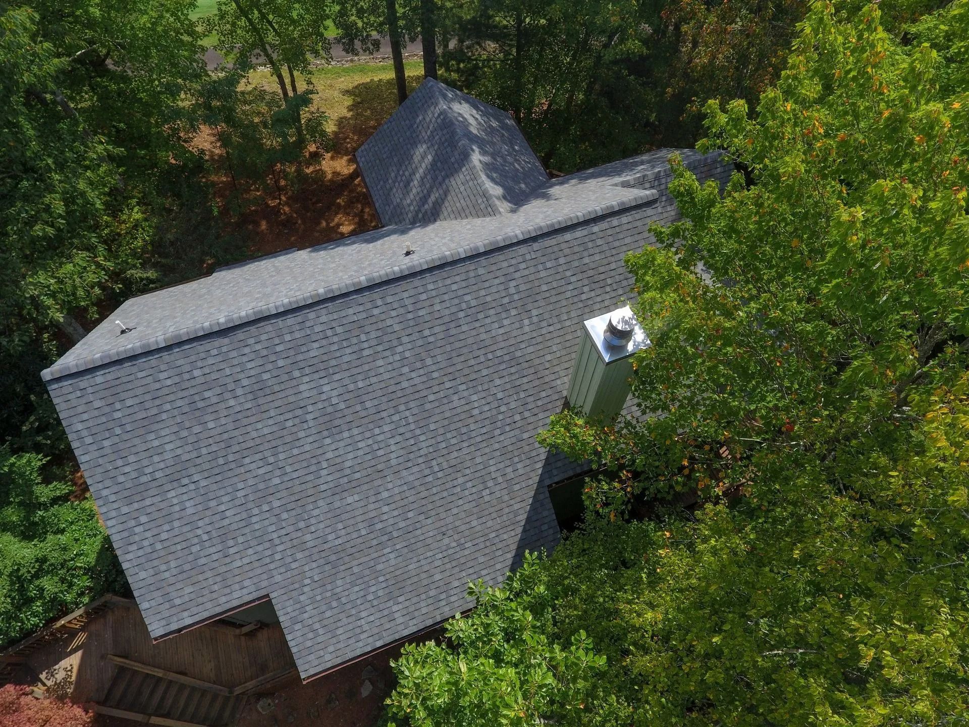 Overhead view of a house with a gray roof, surrounded by green trees. A silver chimney is visible.