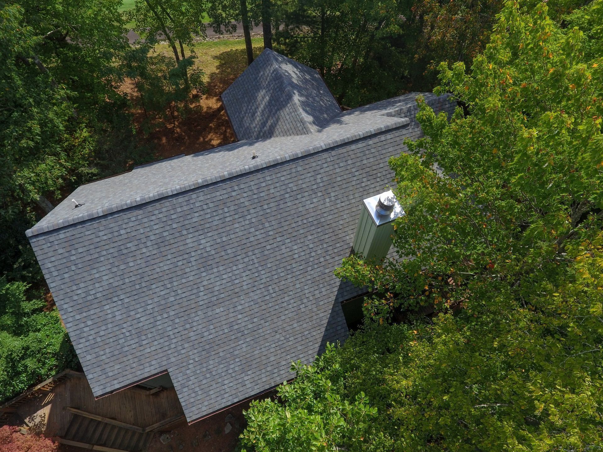Aerial view of a gray shingle roof of a house, surrounded by green trees. A silver chimney is visible.
