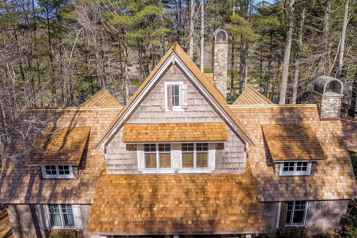 Shingled roof with multiple dormers, chimney. Light brown shingles, gray siding. Surrounded by trees, aerial view.