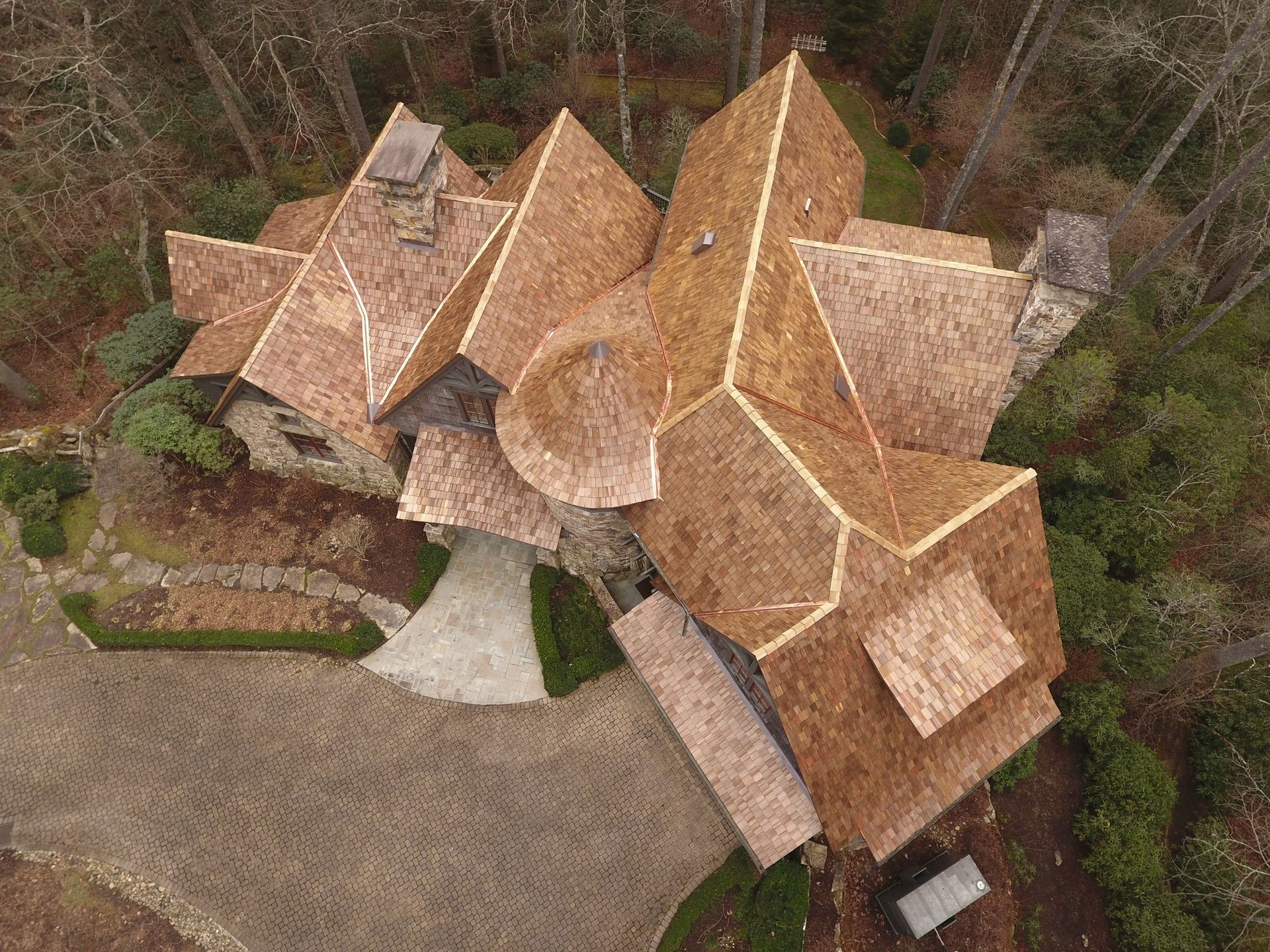 Overhead view of a house with multiple brown, angled roofs. It sits in a wooded area with a gravel driveway.