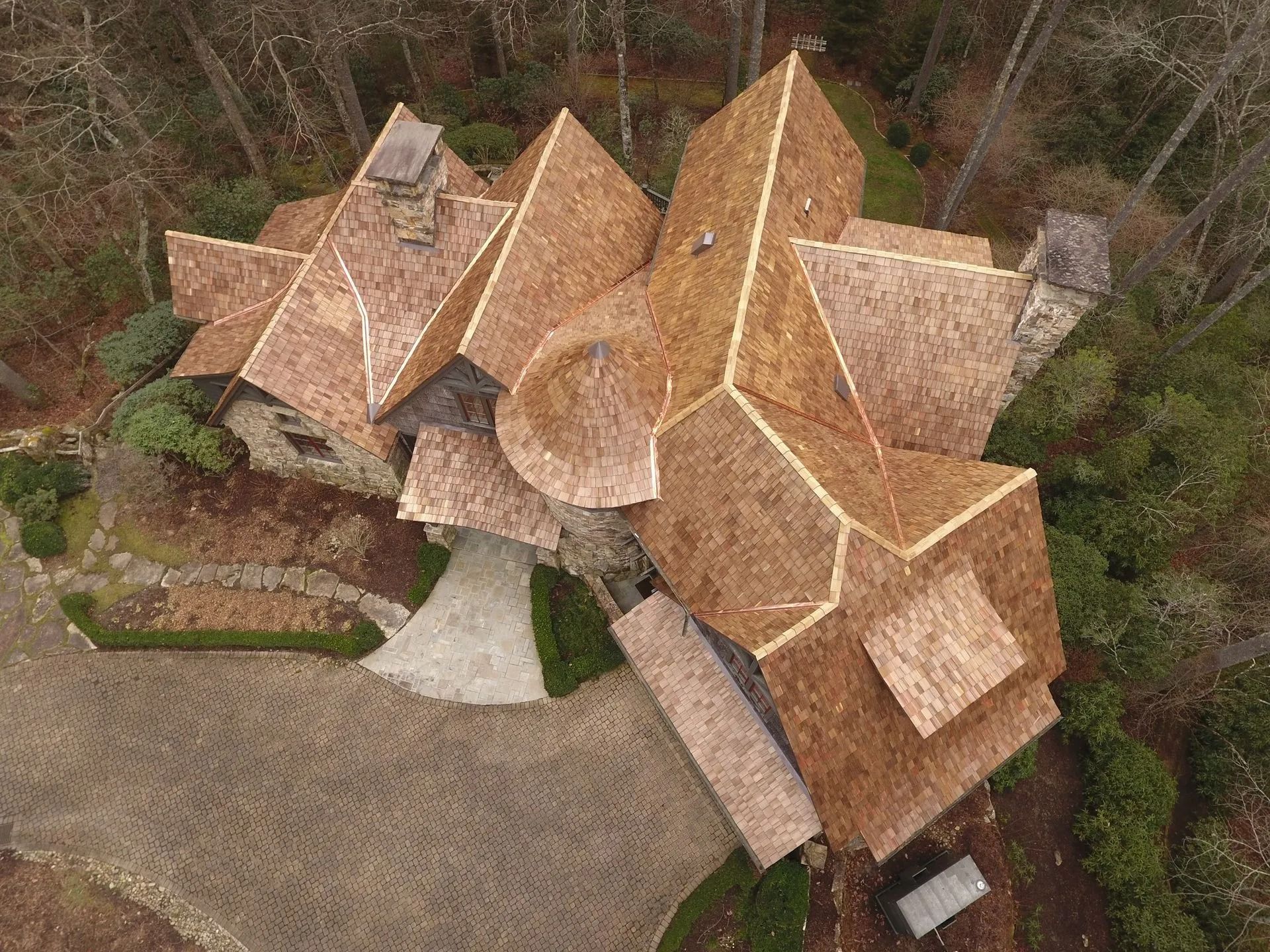 Aerial view of a complex, multi-angled roof house, brown shingles, surrounded by trees and a gravel driveway.