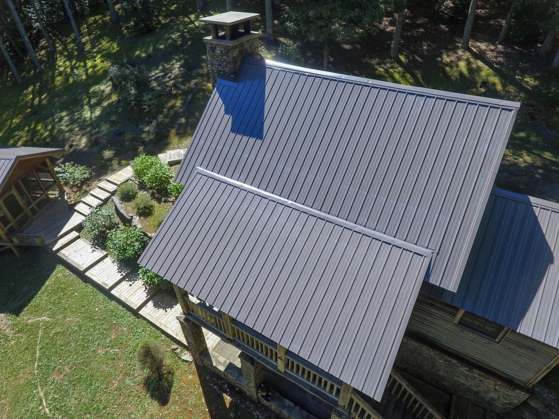 Overhead view of cabin with dark gray metal roof, chimney, stone walkway, and surrounding trees.