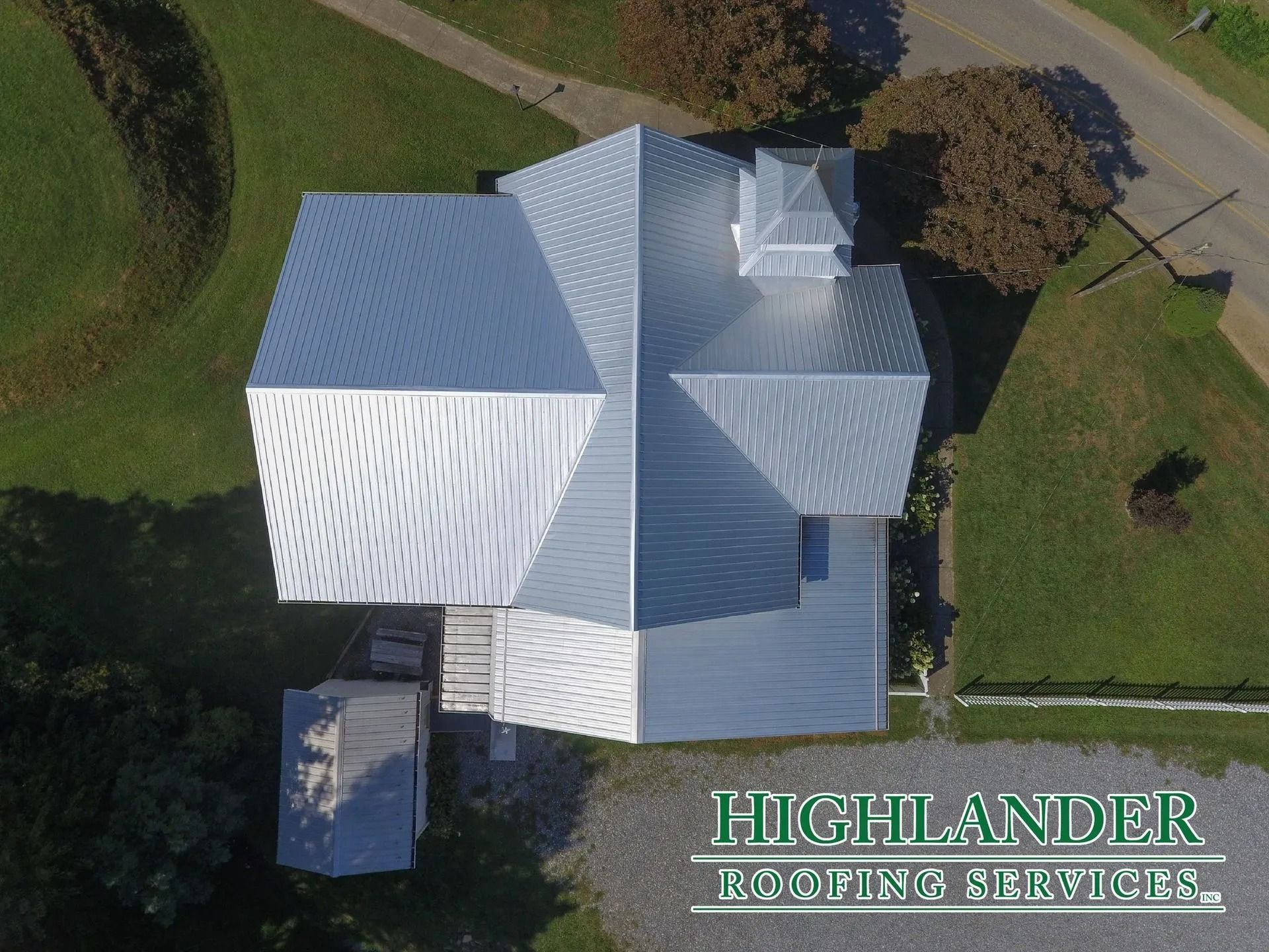 Aerial view of a building with a shiny, silver metal roof. Surrounded by green grass and a gravel driveway.