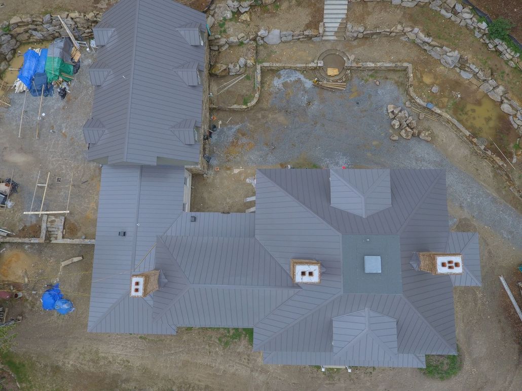 Overhead view of a house under construction with a gray roof and brick chimneys, set in a dirt lot.