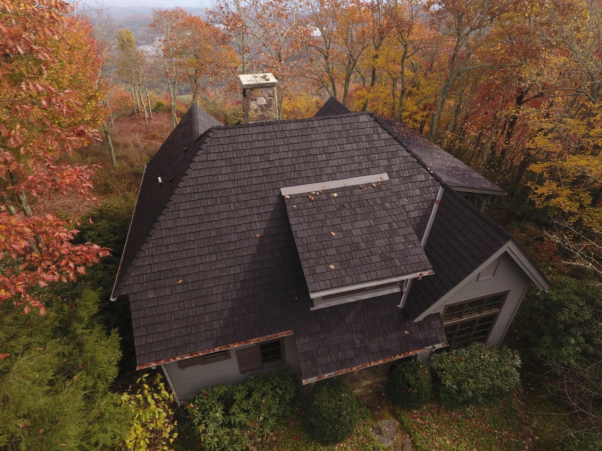Overhead view of a house with a complex, dark-brown shingled roof, surrounded by fall foliage.