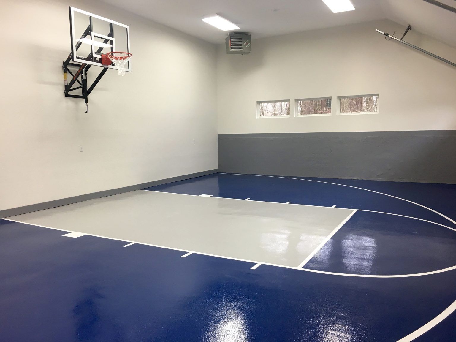 Indoor basketball court with blue and gray flooring, a hoop, and white walls.