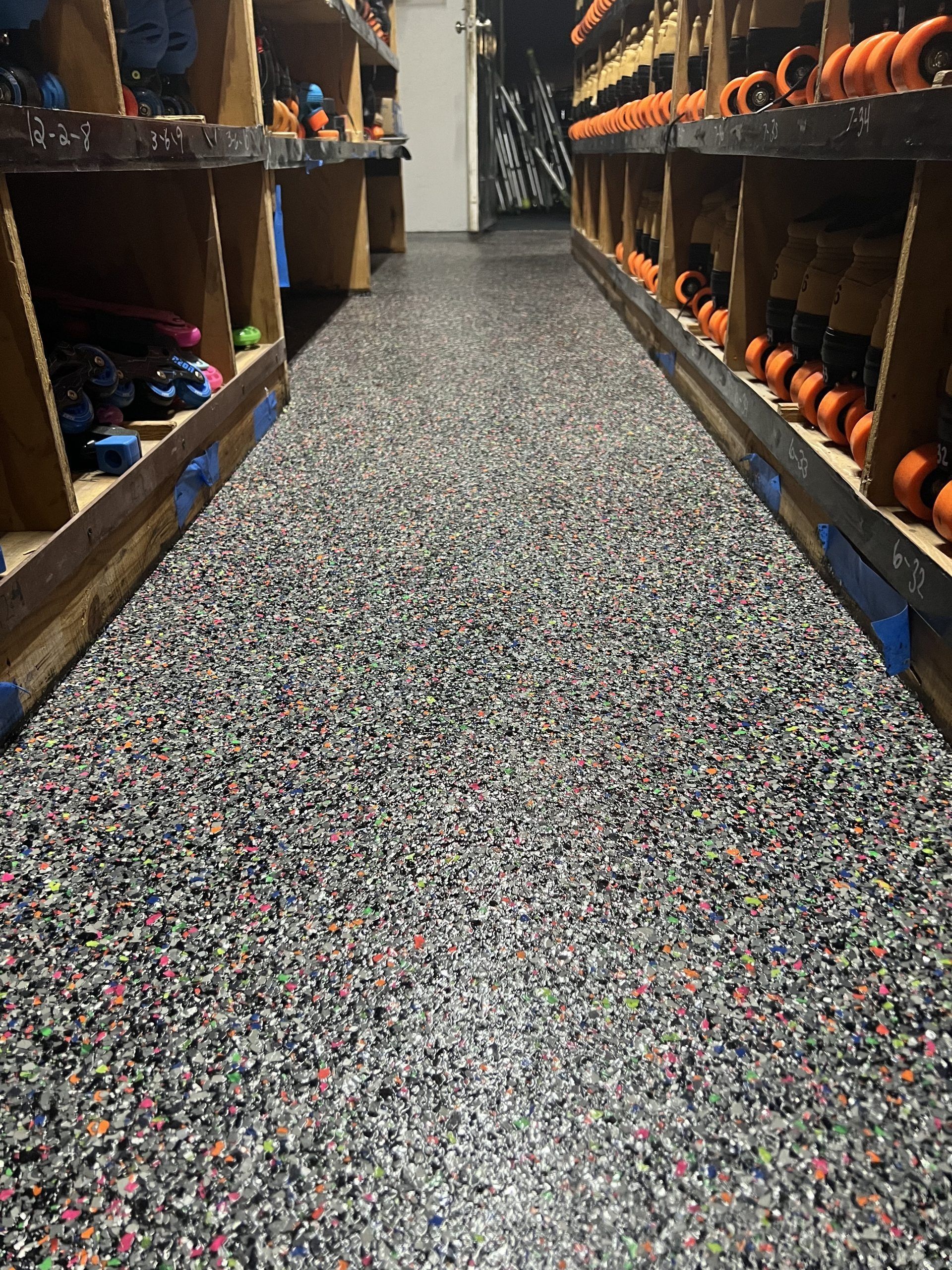 A narrow aisle in a roller skate rental area with shelves of skates on either side; speckled flooring.