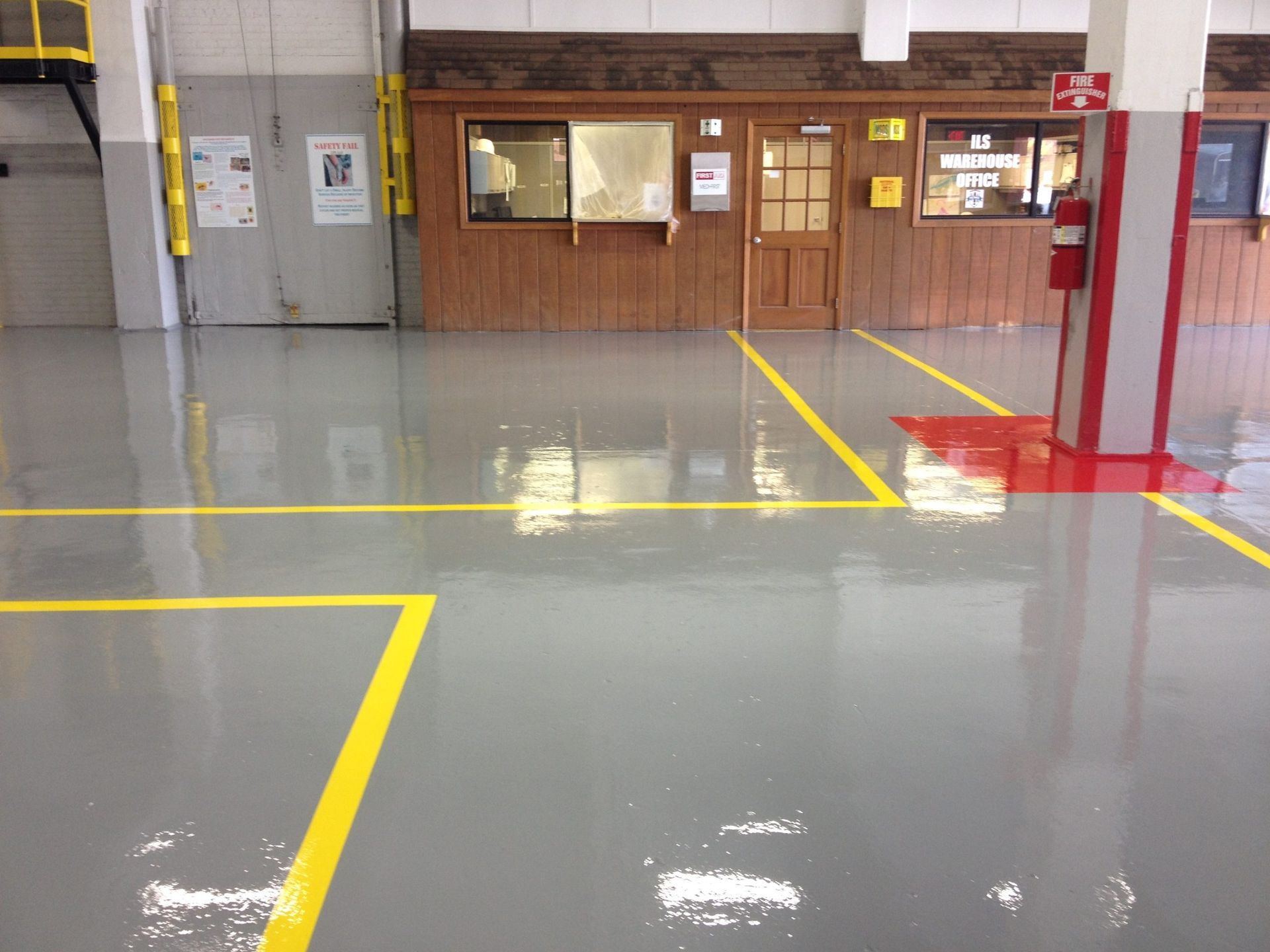 Gray epoxy floor with yellow lines in a shop setting; a wooden wall with a door and windows is in the background.