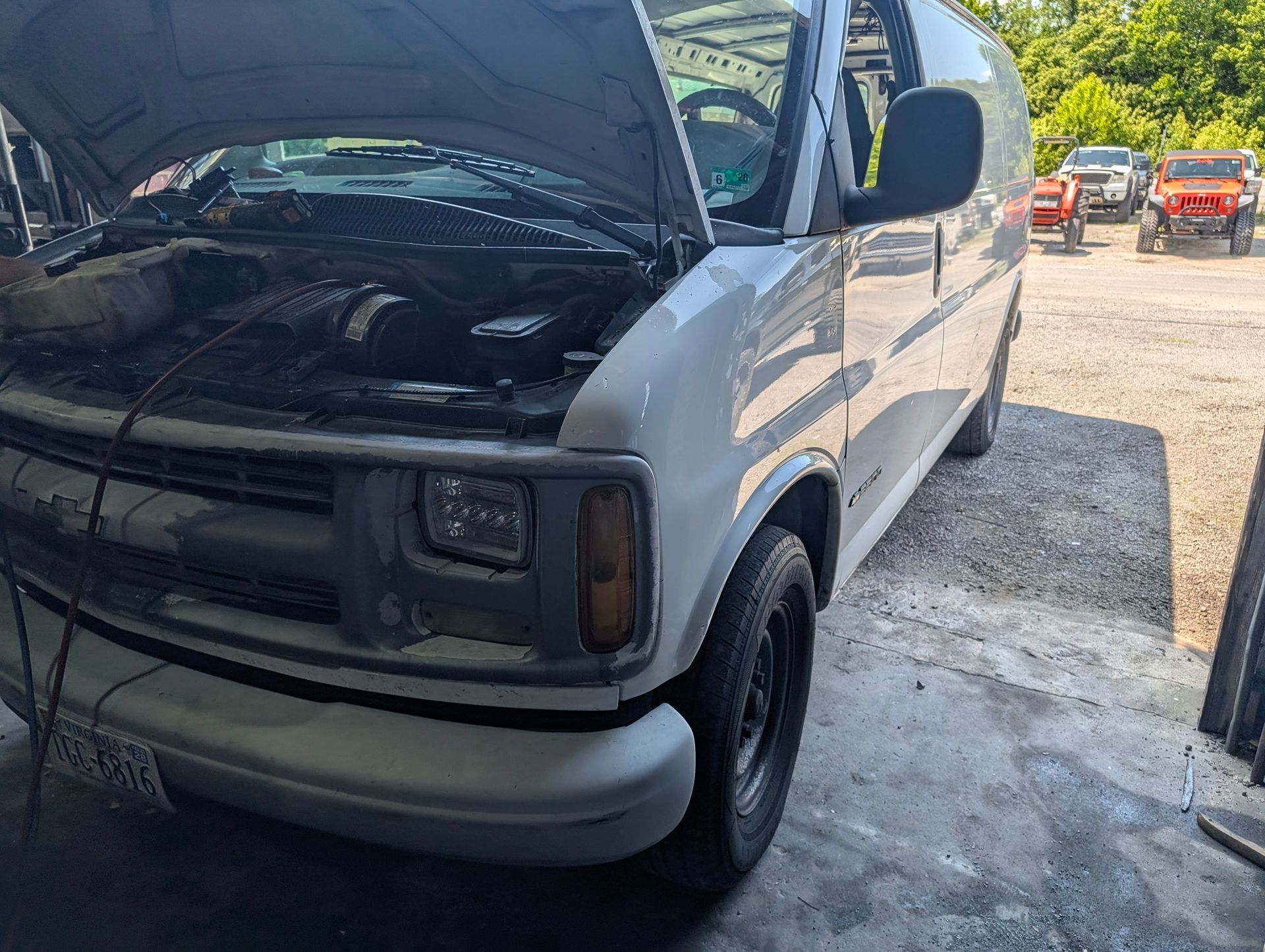 Silver van with open hood in a gravel lot, front end partially disassembled, other vehicles in background
