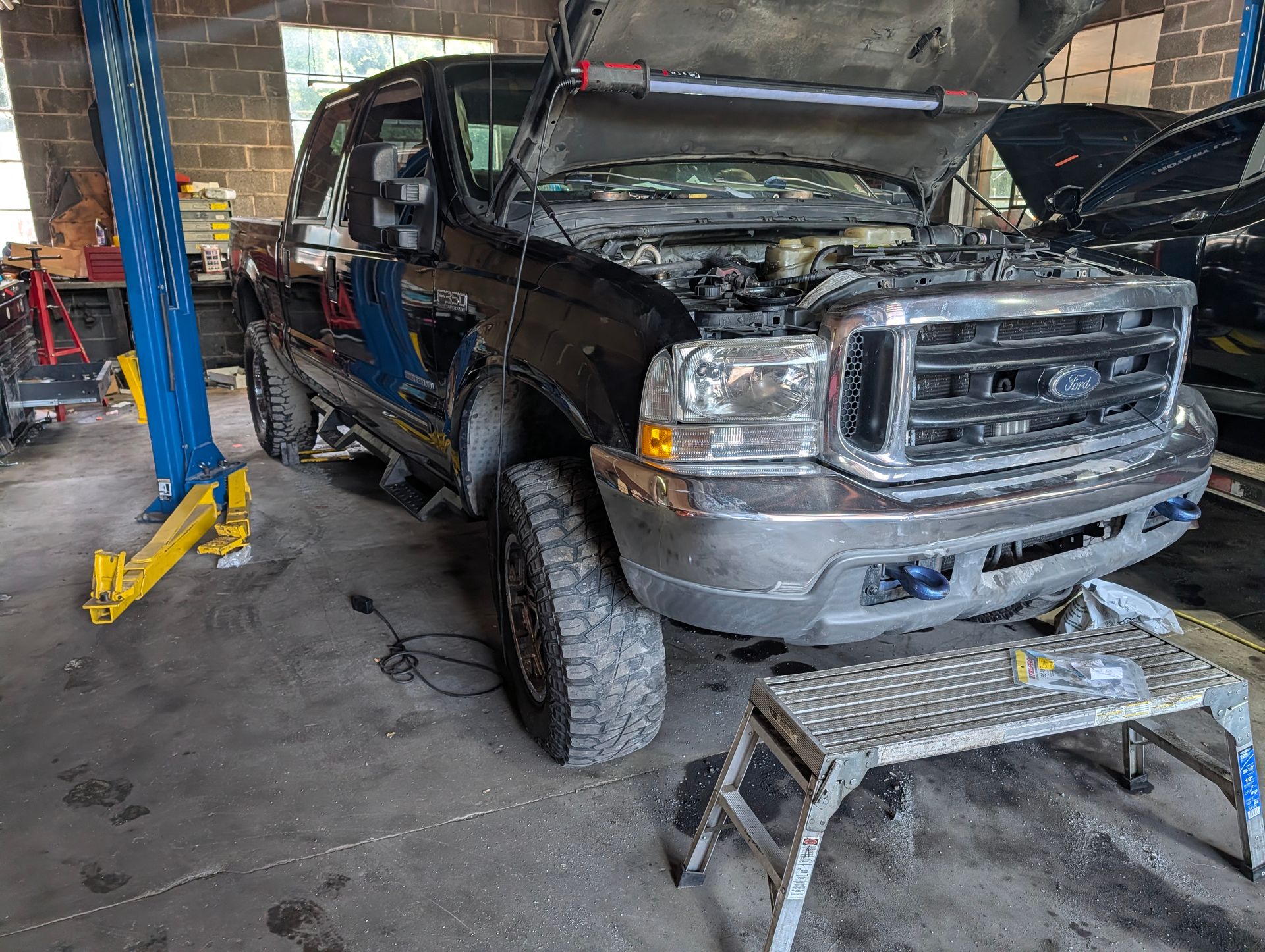 Dark pickup truck with its hood open in a garage, front end raised on a lift stand.