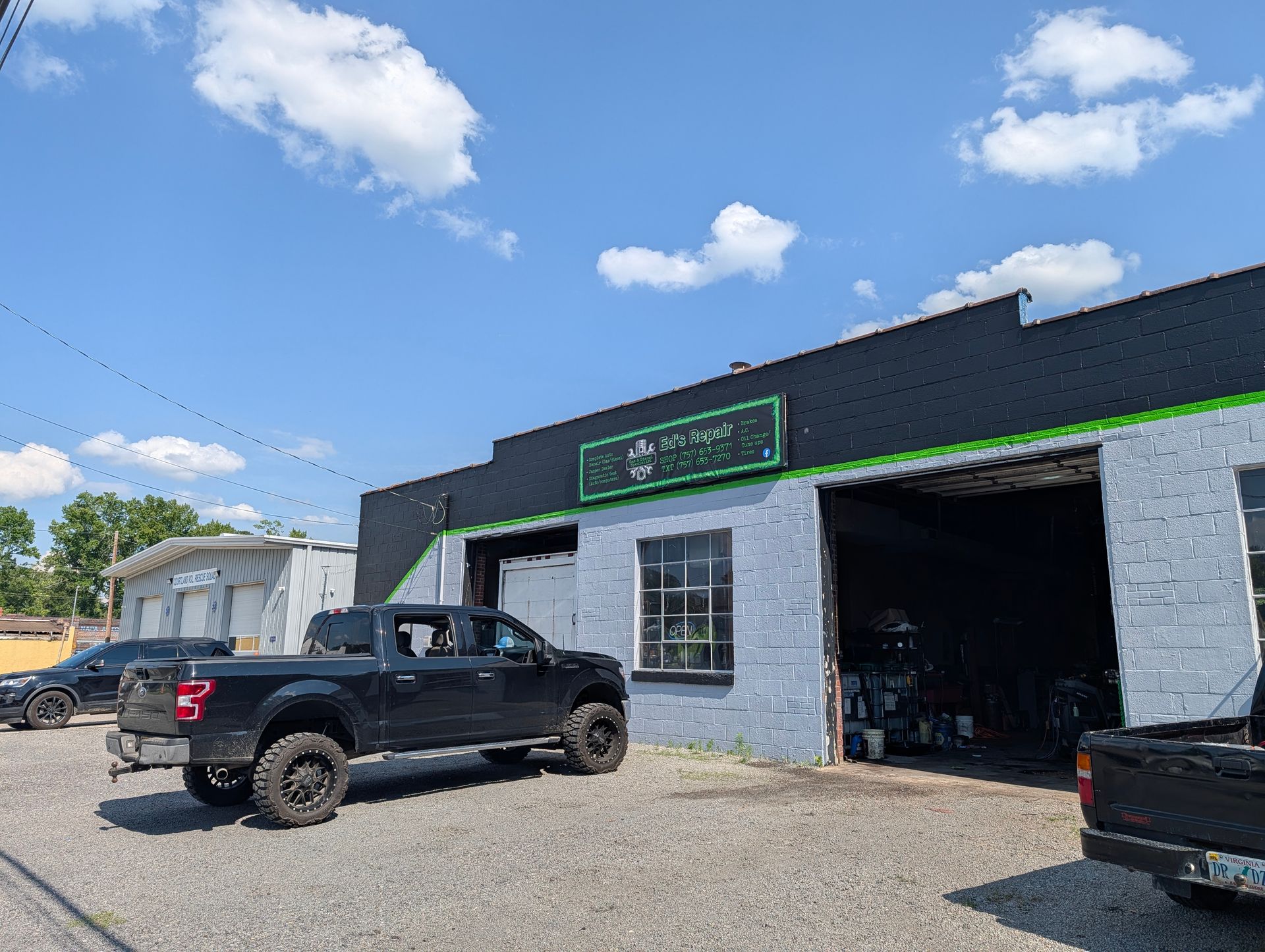 Black pickup trucks outside a gray auto shop with open garage bays under a blue sky