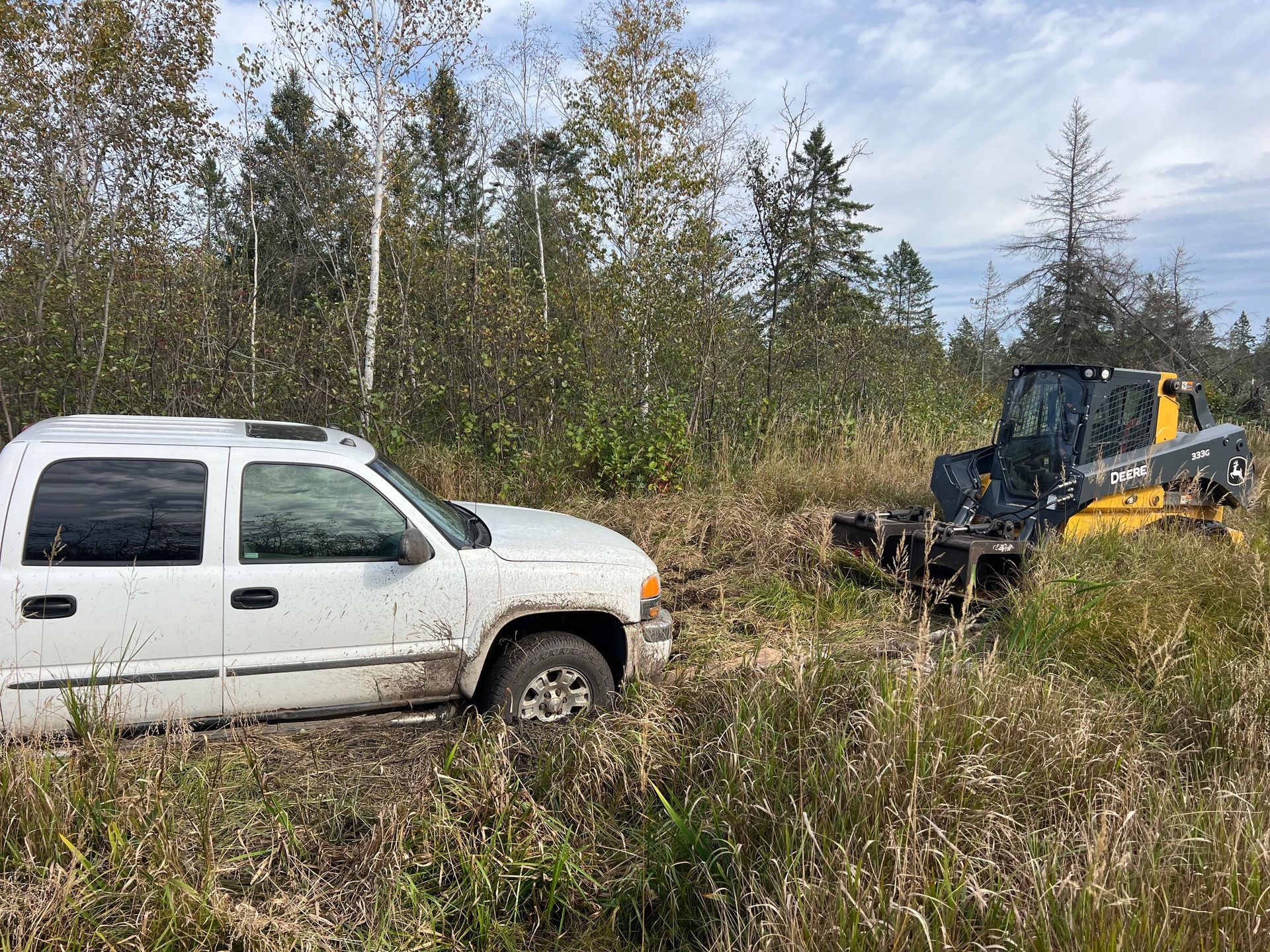 White truck stuck in mud being pulled by a yellow skid steer in a grassy, wooded area.