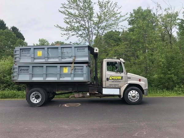 Dump truck on a road, transporting two gray bins. Green trees in the background, overcast sky.