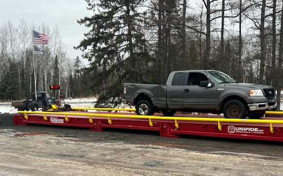 White crane lifting red beams from a flatbed trailer in a snowy outdoor setting.