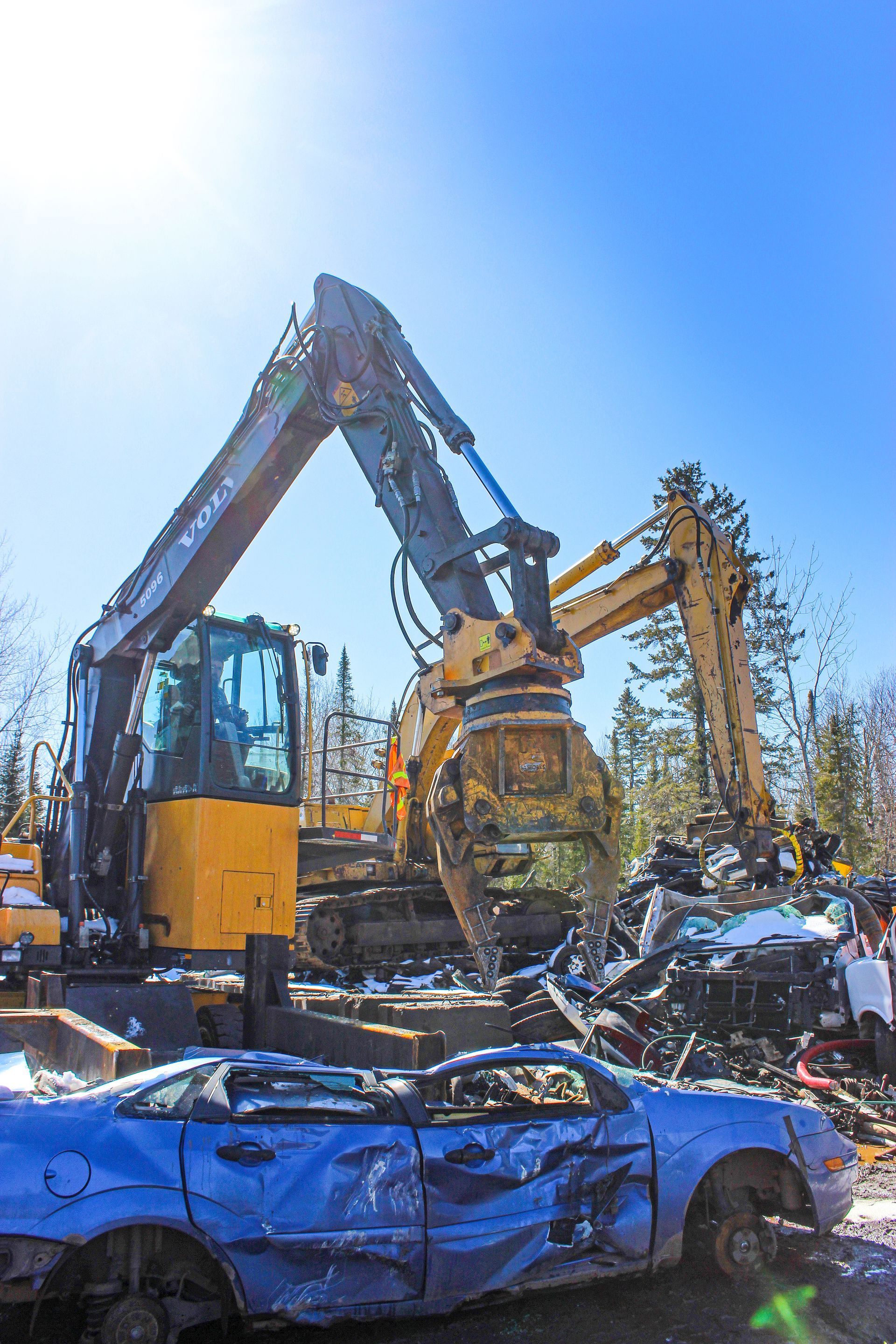 A blue car crushed by a yellow excavator with a large claw, amidst a scrap yard under a bright blue sky.