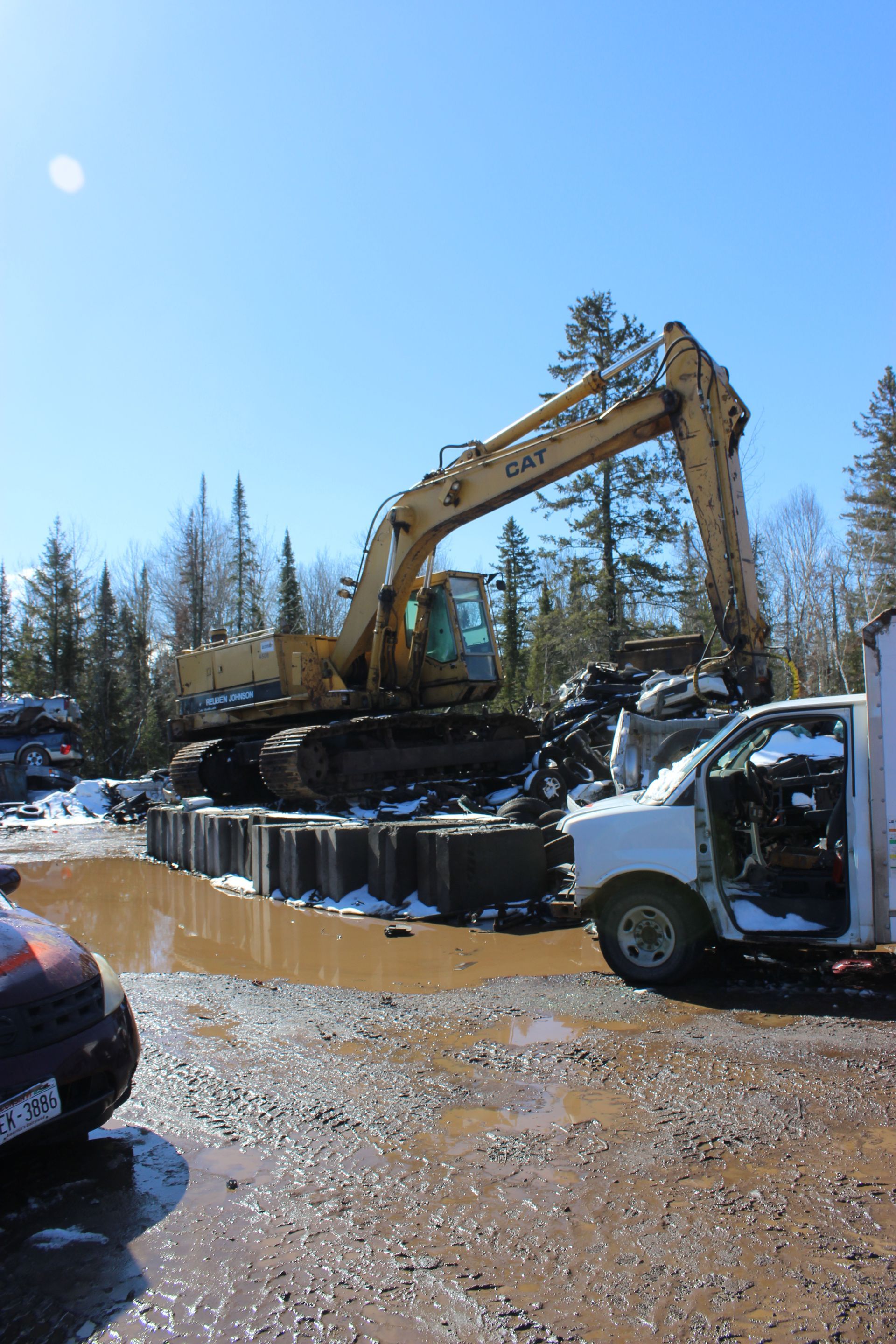 A large yellow excavator at a junkyard, blue sky, muddy ground, with snow present.