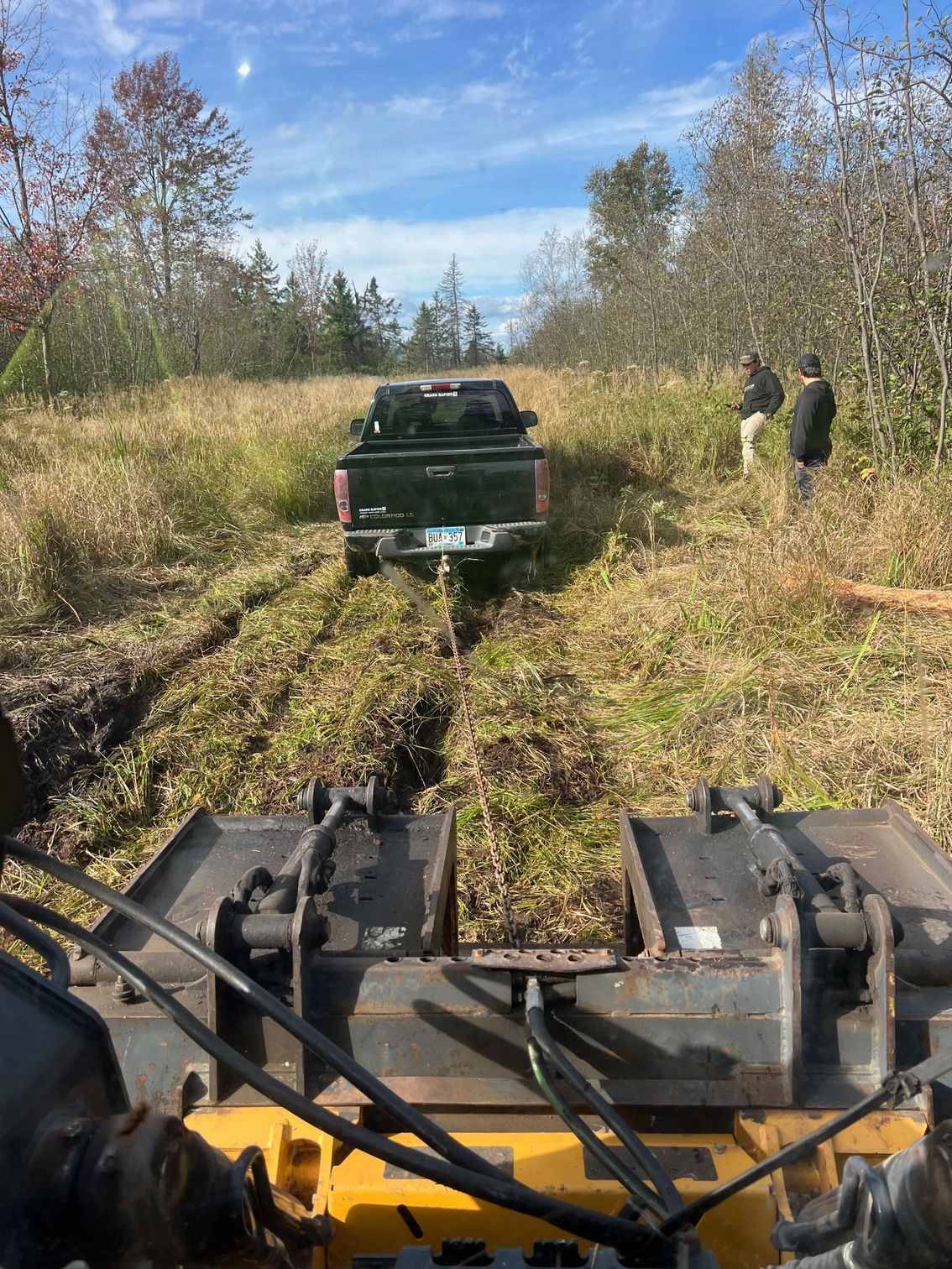 A pickup truck stuck in mud is being pulled out by a front-end loader in a grassy field. Two men watch.