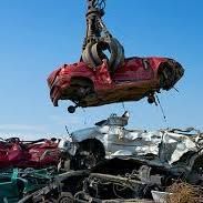 A hydraulic claw lifting a crushed red car over a pile of scrap vehicles at a junkyard under a blue sky.