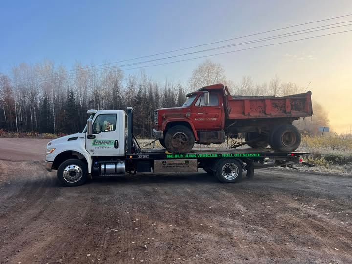 Tow truck carrying a red dump truck on a dirt road, trees in the background.