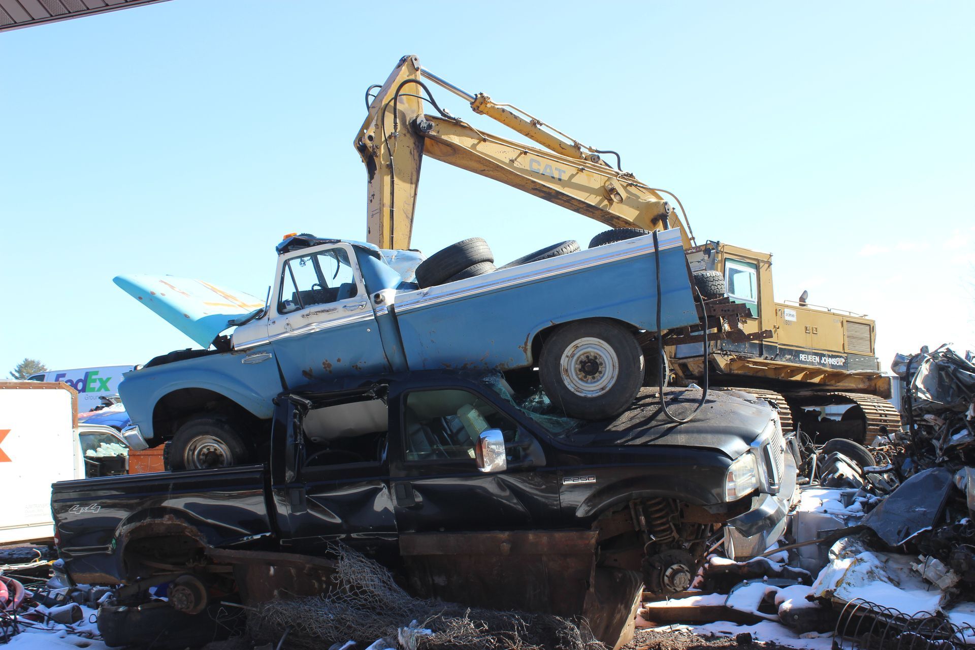 Blue and white pickup truck crushed on top of a black pickup truck in a junkyard with an excavator.