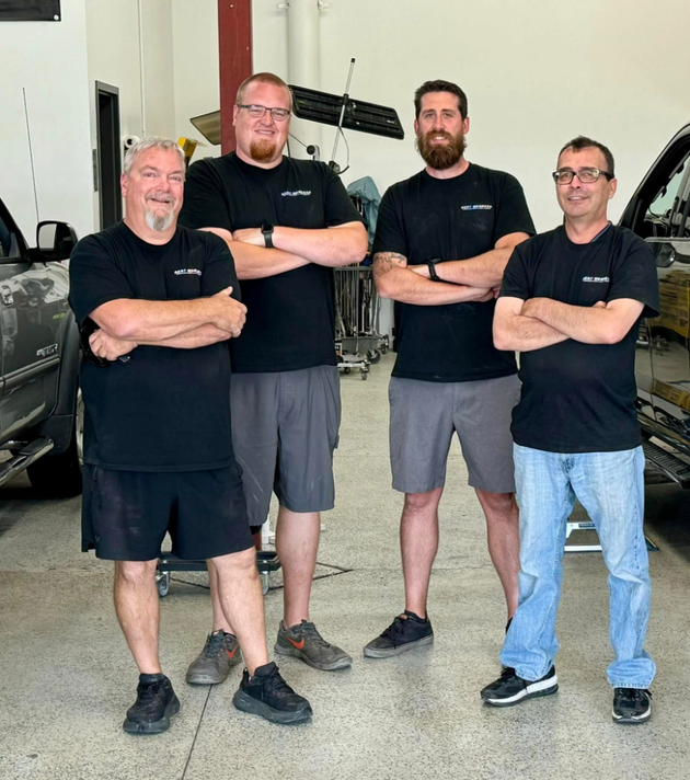Four men in black shirts and shorts/jeans, arms crossed, pose in a repair shop.