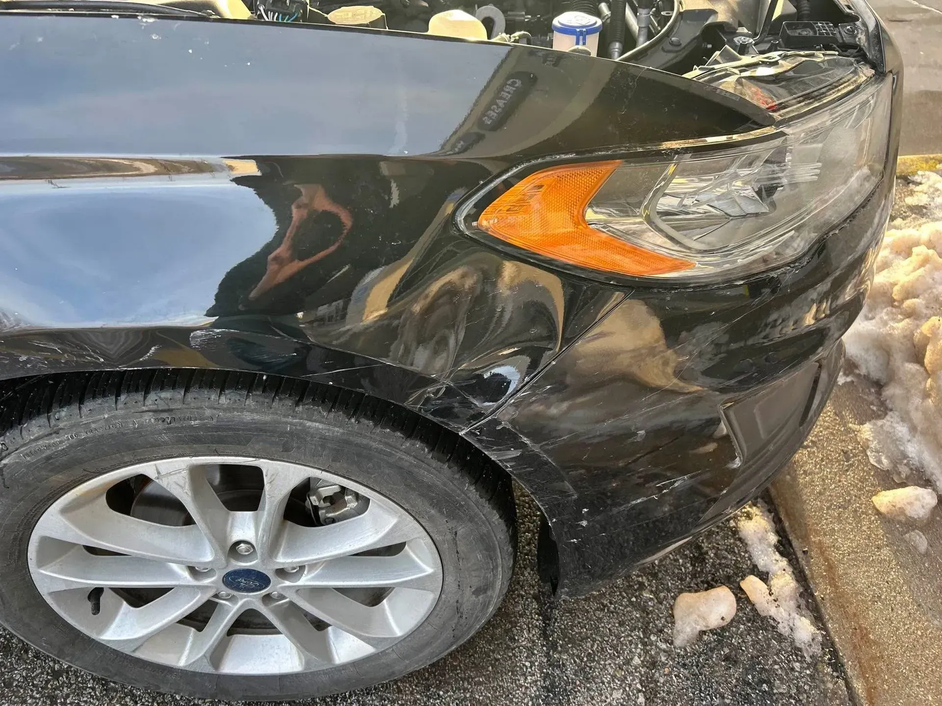 Black Ford car with front-end damage, snow on the ground, and a parking lot.