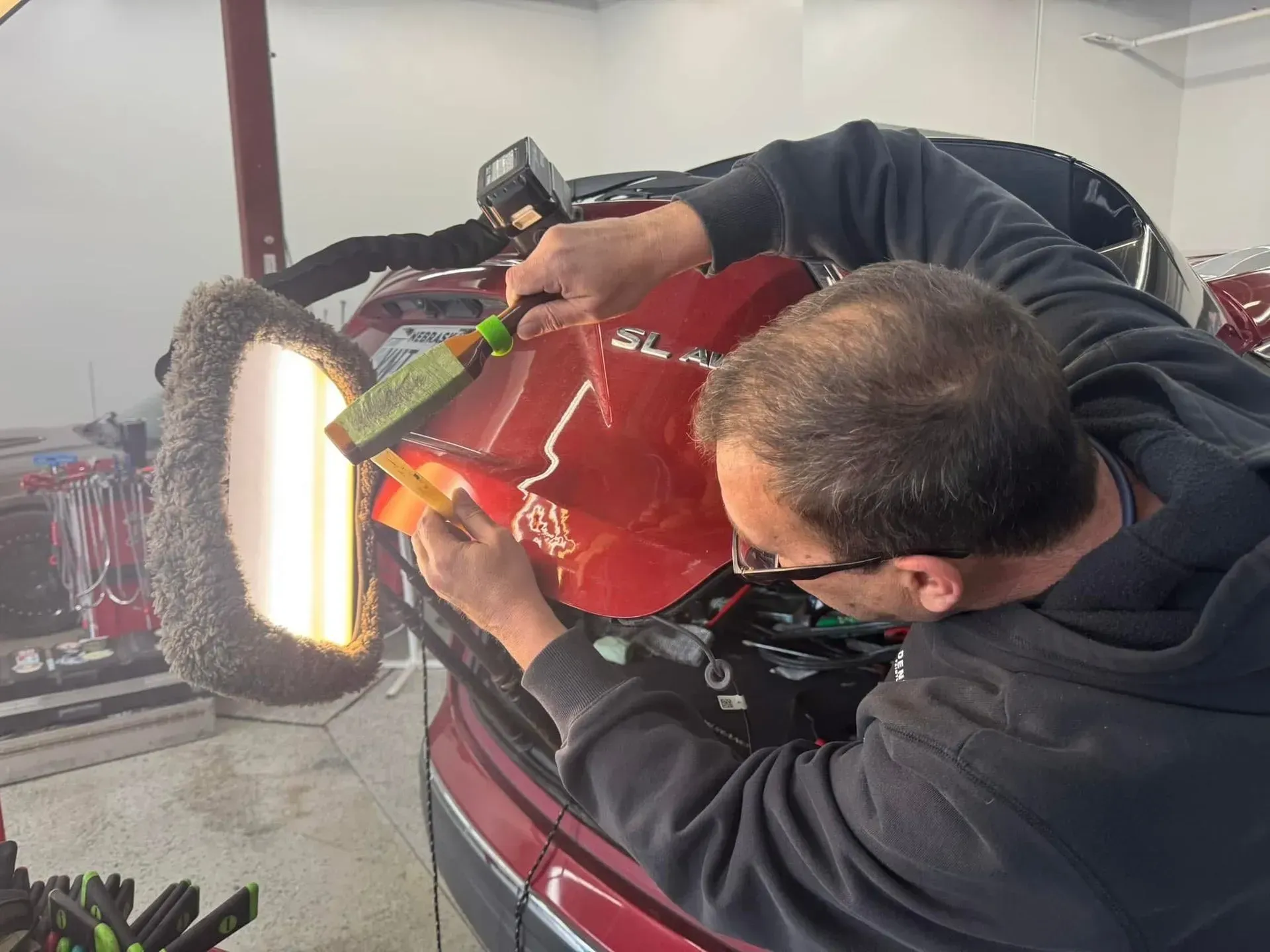A person repairing a red car dent, using tools and a lamp in a workshop.