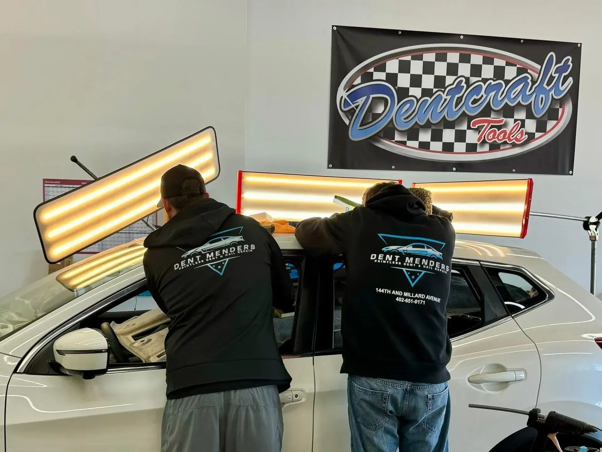 Two people in black hoodies work on a white car using dent removal tools under bright lights.