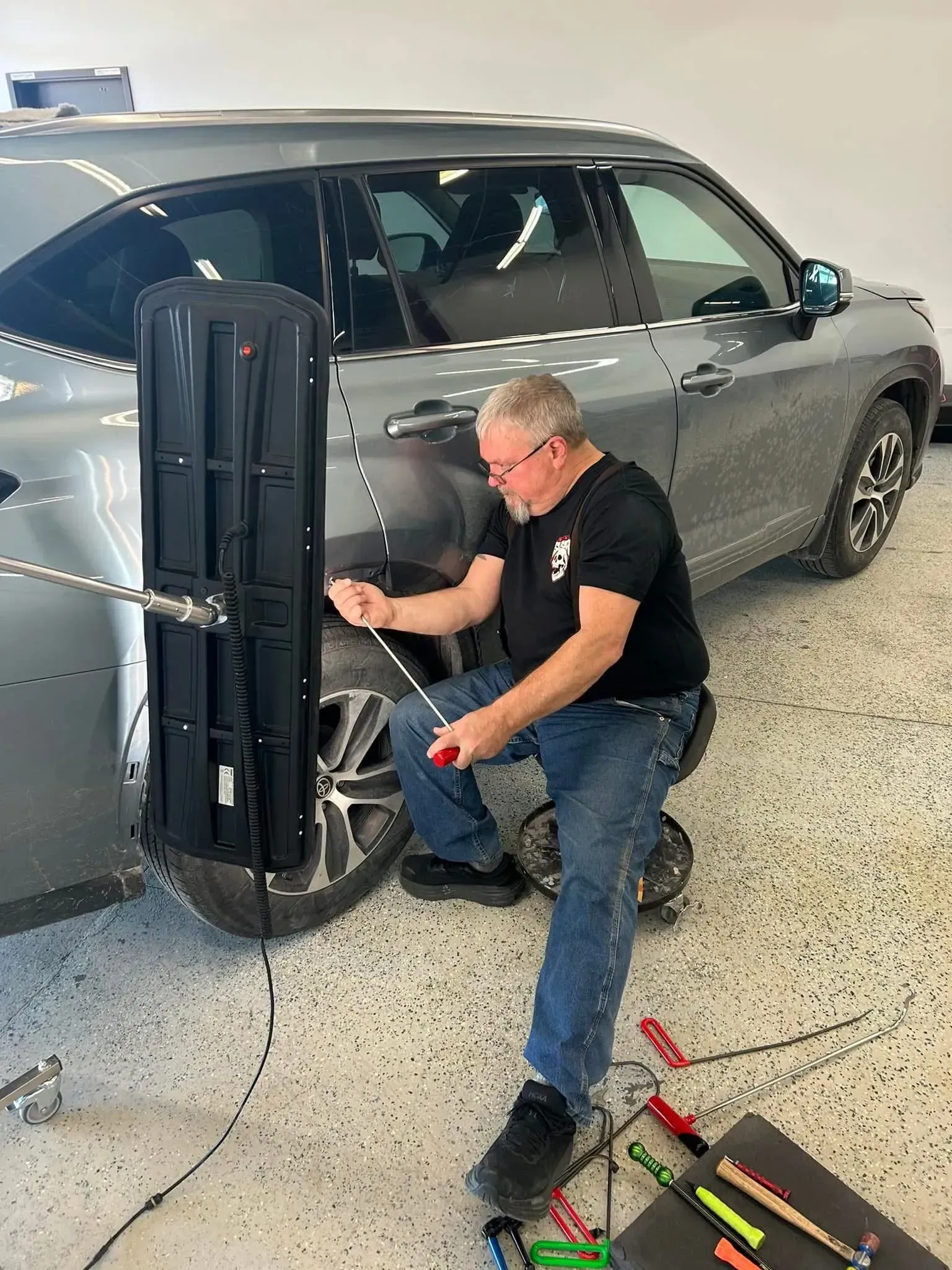 A man repairs a car dent with tools in a garage. Gray car and black tools.