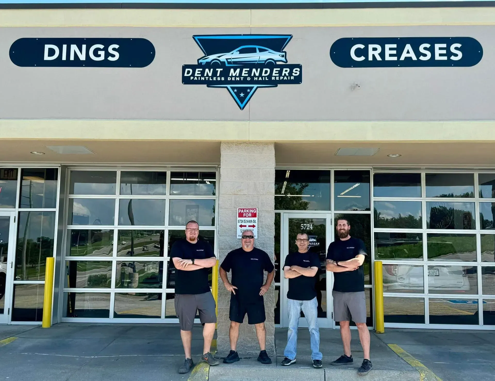 Four men stand in front of "Dent Menders" shop, arms crossed. Black shirts, gray/blue shorts/jeans. Sign above reads "Dings Creases".