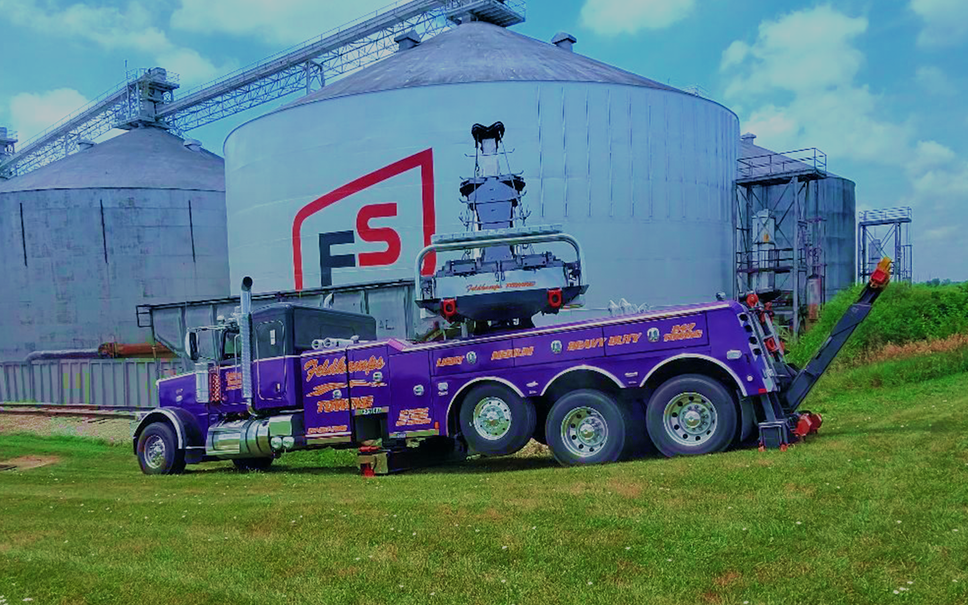Purple tow truck lifting a car in front of large grain silos with the FS logo on the side.