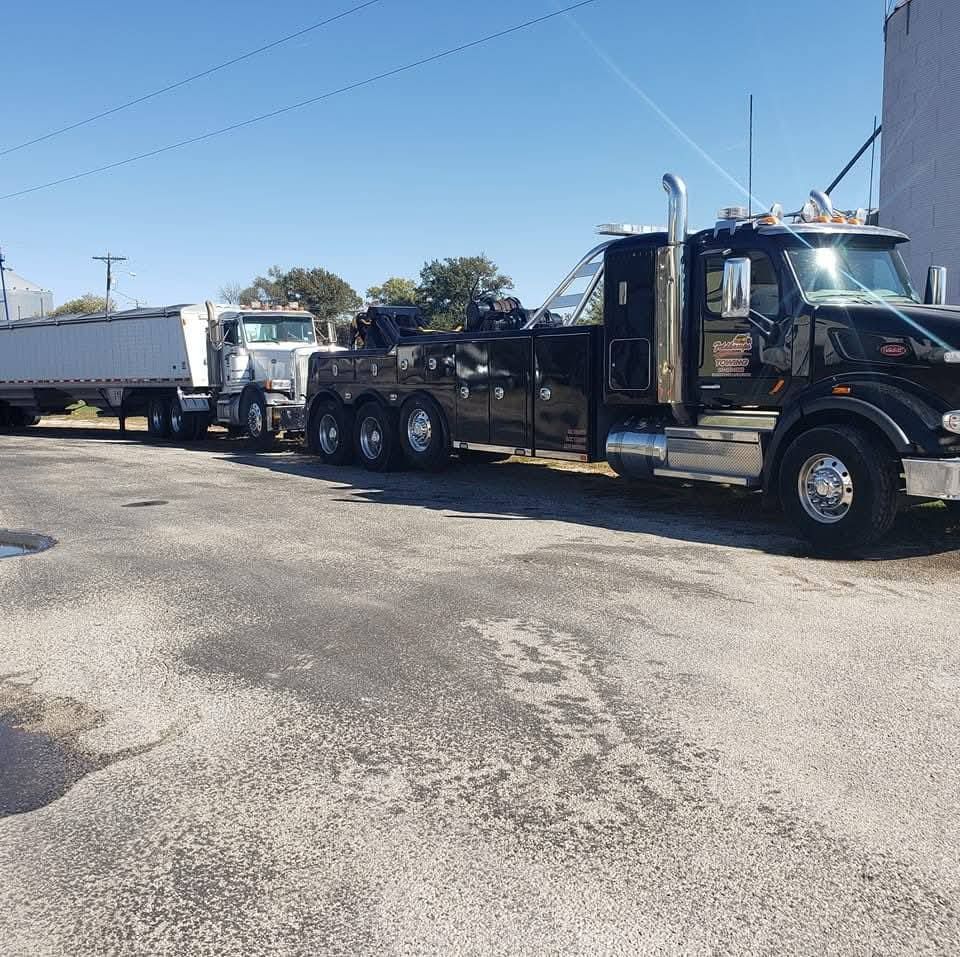 Black tow truck pulling a white semi-truck with trailer on a paved lot under a bright blue sky.