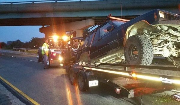 A black Jeep being towed after an accident; on a flatbed trailer under an overpass at night.